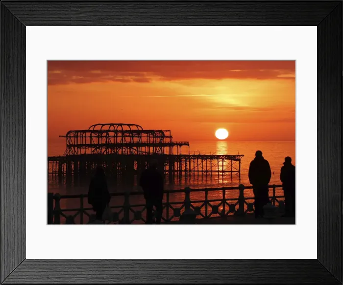 Framed Print : Visitors watch the sun set behind the remains of the West Pier on the seafront at Brighton