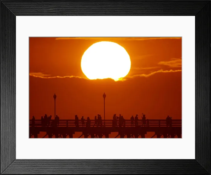 Framed Print : The sun sets behind a cloud as people cool off with a walk along an ocean pier in