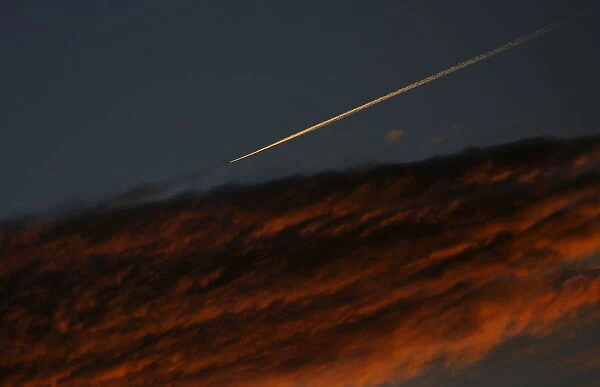 An airplane leaves a vapor trail as it flies past a cloud during dusk as seen