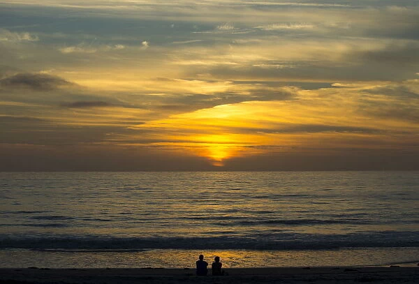 A couple sit on the sand watching the sun set on the Pacific Ocean in Cardiff