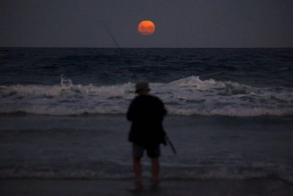 A fisherman prepares to cast his line standing in the surf as a full moon rises at