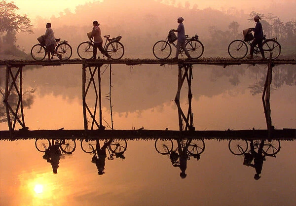 Indonesian villagers push their bicycles across a bamboo bridge as sun rises behind them outside