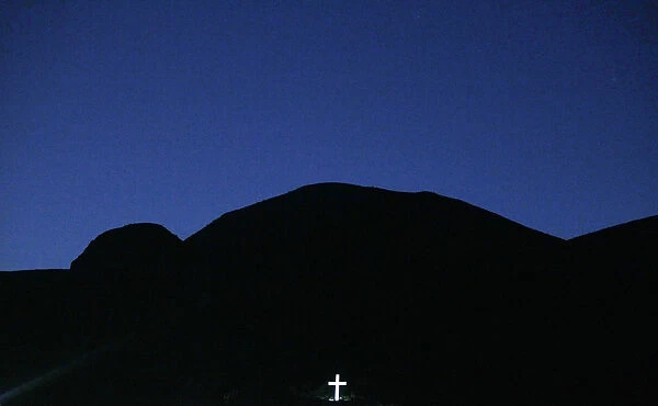 A large crucifix is illuminated below the mountains as the sun sets in the Peloponnese