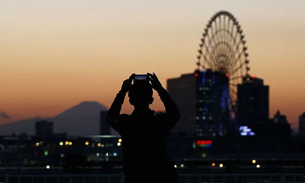 A man takes a picture of a ferris wheel and Mount Fuji during sunset in Tokyo