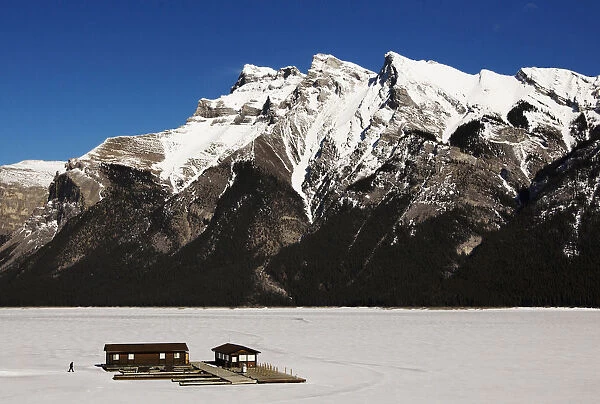 A man walks over a frozen Lake Minnewanka to a boathouse in front of the Canadian