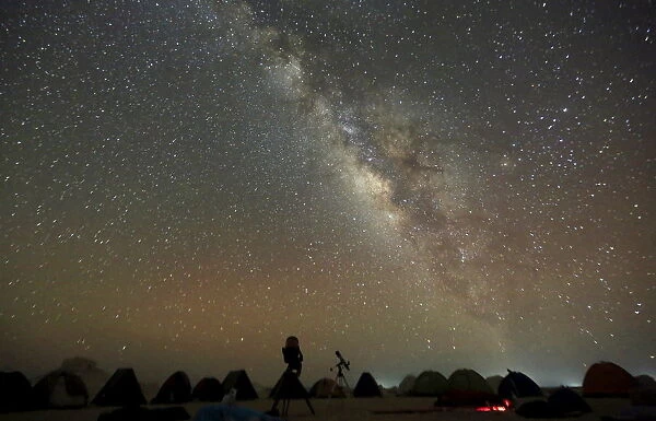 The Milky Way is seen in the night sky around telescopes and camps of people over