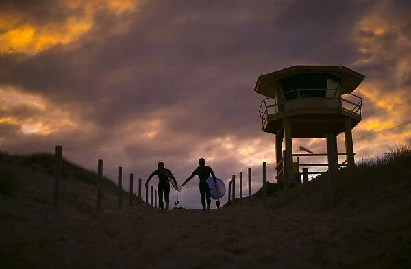 A pair of female surfers march up the beachhead alongside a lifesavers hut at sunset