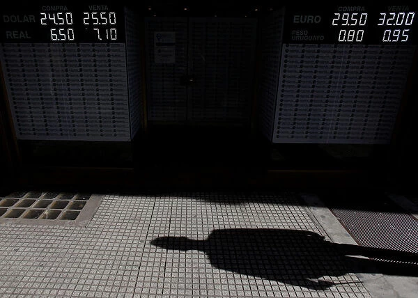 A pedestrians shadow is cast next to an electronic board showing currency exchange