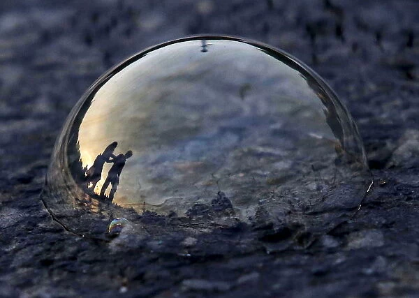 People are reflected on a soap bubble lying on a road during the evening in New Delhi