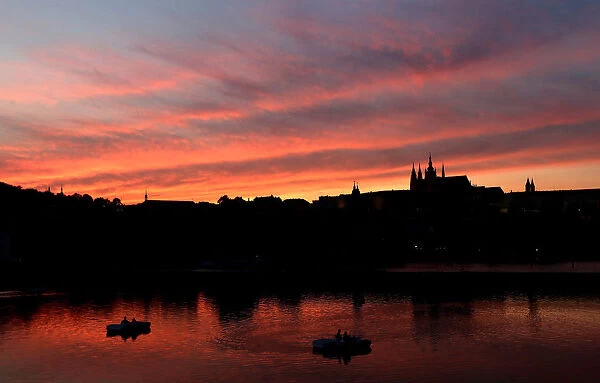 People ride pedal boats on the Vltava River as the sun sets over the Prague Castle in