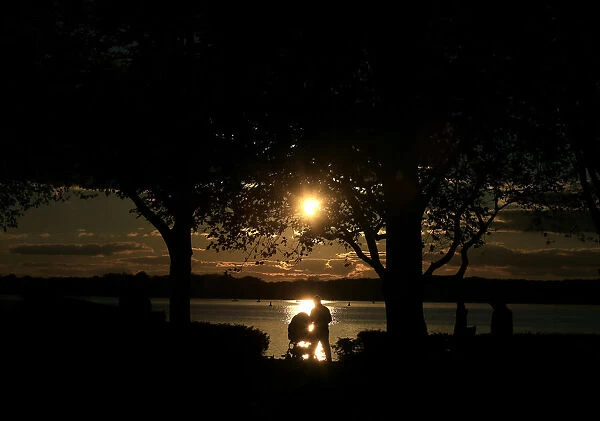 A person pushing a pram is silhouetted against the reflection of the sun in lake Tegel