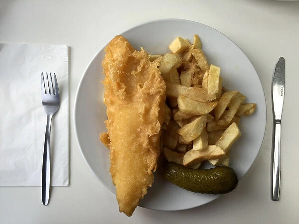A plate of traditional fish and chips with a gherkin on a table in a seafront fish