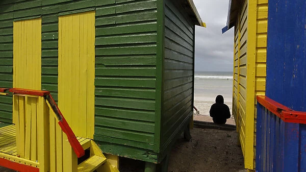 A spectator sits between beach huts as surfers prepare to take to the water in an