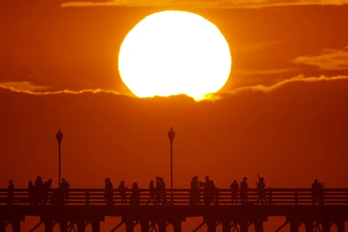 Framed Print : The sun sets behind a cloud as people cool off with a walk along an ocean pier in