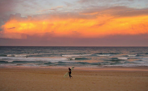 A surfer carrying his board walks out of the ocean as a cloud is lit by the setting