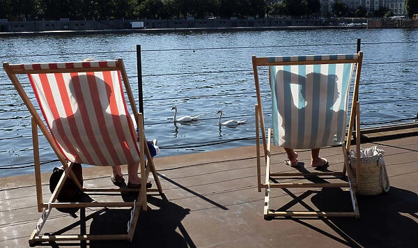 Swans swim on the Vltava River as people sunbathe on deck chairs during a heat wave