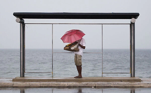 A vendor holds up an umbrella as he seeks shelter from the heavy rain under a bus stand