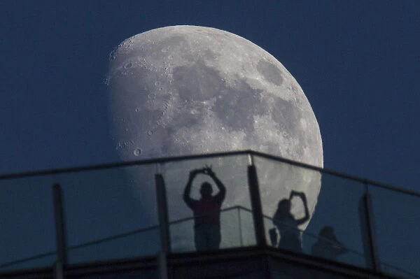 Visitors stand on the roof of a skyscraper as the moon rises over the skyline of Pudong