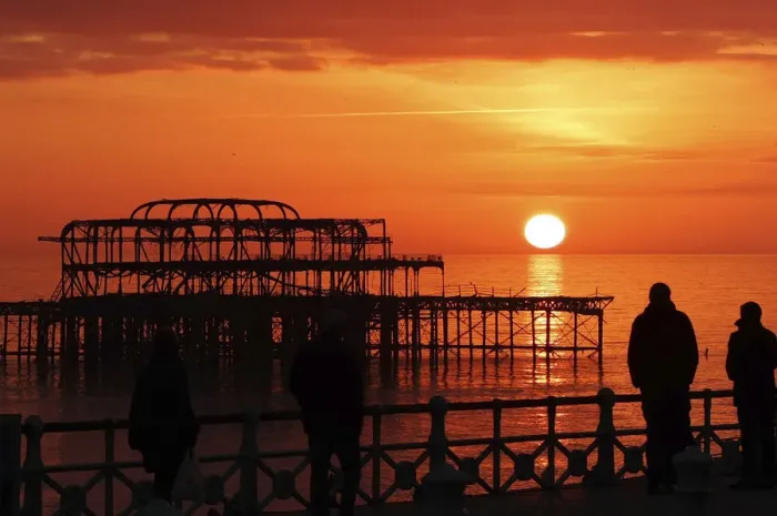 Framed Print : Visitors watch the sun set behind the remains of the West Pier on the seafront at Brighton