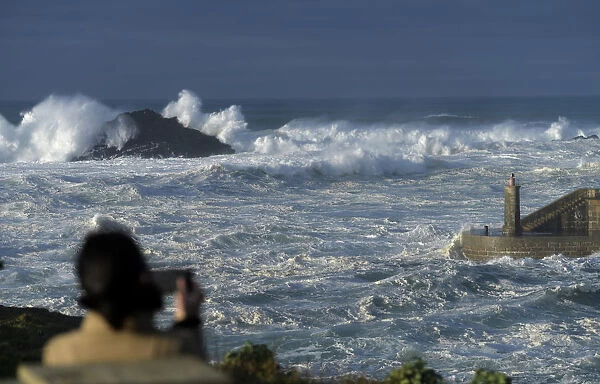 A woman takes pictures of waves as they crash against a lighthouse in the port town of