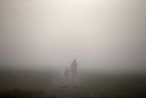 A woman walks with a child through a fog at a field at a makeshift camp for migrants