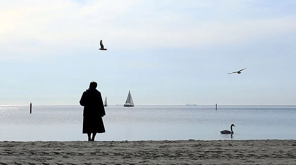 A woman, wrapped up against the cold wind, stands on the beach as she watches a swan