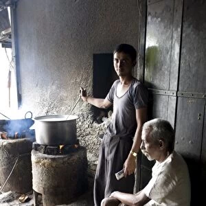 Young man stirring milk for chai, with blind man sitting beside him, roadside chai stall