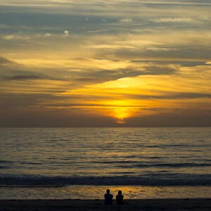 A couple sit on the sand watching the sun set on the Pacific Ocean in Cardiff