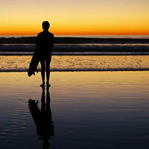 A lone surfer stands watching the waves after surfing as the sun sets in Leucadia