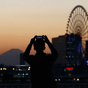 A man takes a picture of a ferris wheel and Mount Fuji during sunset in Tokyo