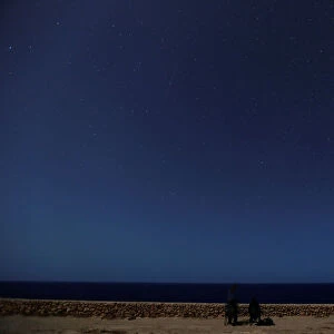 A meteor streaks past stars as people face the sea hoping to catch a glimpse of shooting