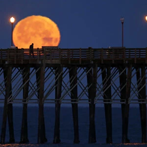 The moon sets in the early morning hours as a man walks along an ocean pier in Oceanside