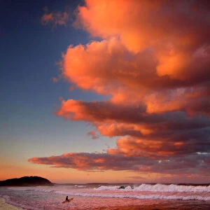 Surfer holding her board wades through the surf as clouds above are lit by the setting