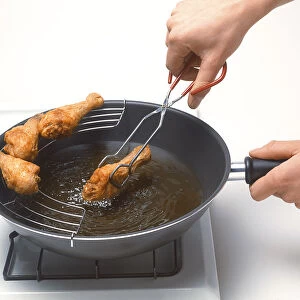 Hands using tongs to move deep-fried chicken drumstick from pan onto draining rack