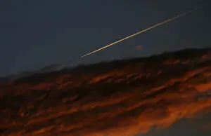 An airplane leaves a vapor trail as it flies past a cloud during dusk as seen