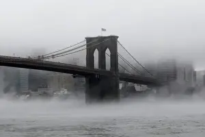 The Brooklyn Bridge is seen partially in fog from in front of the Manhattan skyline