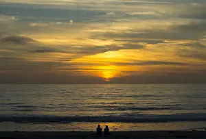 A couple sit on the sand watching the sun set on the Pacific Ocean in Cardiff