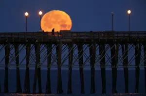 The moon sets in the early morning hours as a man walks along an ocean pier in Oceanside