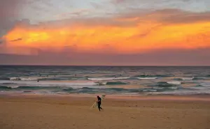 A surfer carrying his board walks out of the ocean as a cloud is lit by the setting