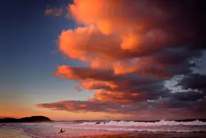 Surfer holding her board wades through the surf as clouds above are lit by the setting