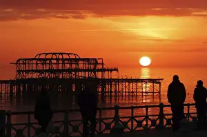 United Kingdom Prints: Visitors watch the sun set behind the remains of the West Pier on the seafront at Brighton